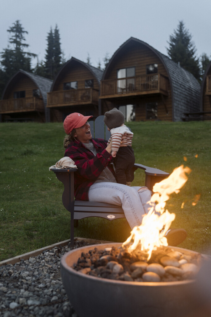 mom and baby sit fireside with cabins in the background